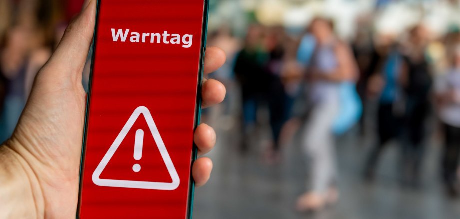 Eine Hand hält ein Smartphone, auf dessen Bildschirm ein roter Warnhinweis mit einem weißen Ausrufezeichen und dem Text 'Warntag' angezeigt wird. Im Hintergrund ist eine unscharfe Menschenmenge in einer belebten Umgebung zu sehen.