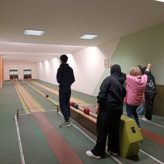 Das Bild zeigt mehrere Personen auf einer Kegelbahn. Eine Person steht am Anfang der Bahn und wartet, während andere auf der rechten Seite Kegelkugeln werfen. Es sind mehrere Kegelbahnen zu sehen, und die Kegel sind im Hintergrund aufgestellt. Eine Person trägt eine pinke Oberbekleidung, während andere dunkle Kleidung tragen. Die Beleuchtung erfolgt durch Deckenlampen, und die Wände sind grün gestrichen. Ein Gemälde hängt an der rechten Wand.