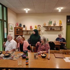 Das Bild zeigt eine Gruppe von fünf Personen, die um einen Tisch sitzen. Auf dem Tisch befinden sich Snacks, Getränke und Kerzen. Im Hintergrund sind Regale mit verschiedenen dekorativen Objekten und Trophäen an der Wand angebracht. Die Personen wirken in Gespräche vertieft, und eine Person im Hintergrund nutzt ein Smartphone. Der Raum ist schlicht eingerichtet und wird von warmem Licht beleuchtet.