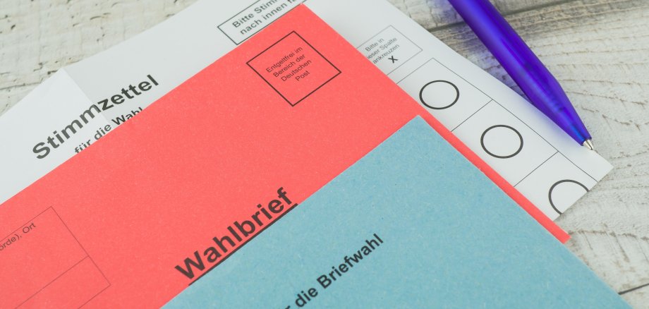 Briefwahlunterlagen bestehend aus einem Wahlbriefumschlag, einem Stimmzettel und einem blauen Kugelschreiber, auf einer hellen Holzoberfläche liegend.