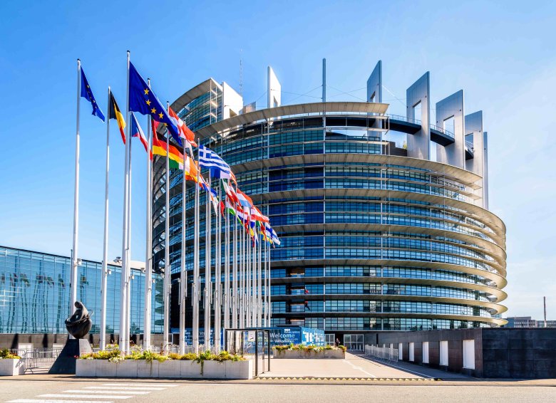 Das Europaparlament in Straßburg, eine moderne, halbkreisförmige Glasfassade mit mehreren Stockwerken. Vor dem Gebäude wehen Flaggen der Mitgliedsstaaten der Europäischen Union sowie die EU-Flagge. Im Vordergrund steht eine Skulptur, und ein Schild mit der Aufschrift 'Welcome' ist am Eingangsbereich zu sehen. Der Himmel ist klar und blau.